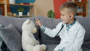boy dressed as a doctor, pretending to examine a white teddy bear using a toy stethoscope
